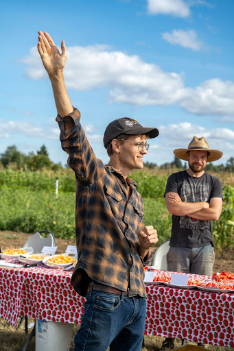 Facilitator speaking to participants at an outdoor dry farming field day, with another participant standing by tables of produce in the background.