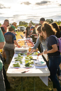 People gathered around long outdoor tables sampling sliced cucumbers and other produce during a field day.