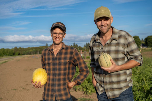 Two people standing in a field holding dry‑farmed melons and smiling toward the camera.